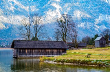 Dağlarda güzel göl, yalnız bank, arka planda ahşap bir ev, evlerin arkasında güzel ağaçlar ve çamlarla kaplı dağlar vardır. Gündüz