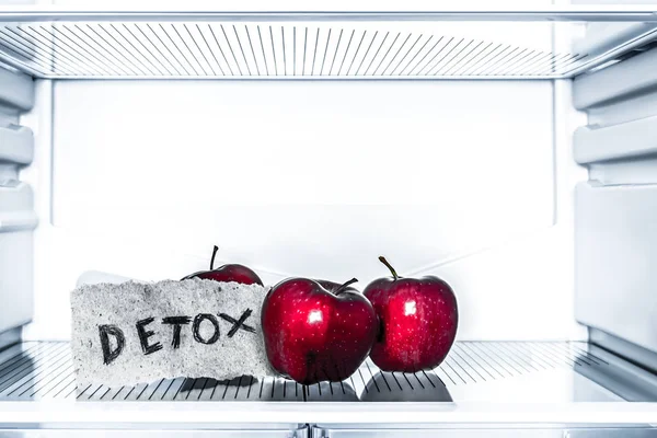 Three red apples lie on an empty shelf of the refrigerator. A piece of paper with the word detox. Diet and weight loss concept. Close Up
