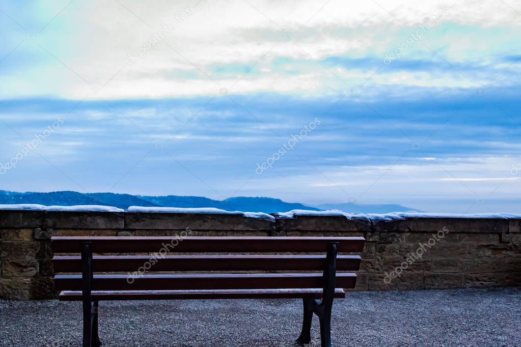 High in the mountains, in the courtyard of Hohenzollern castle is a lonely bench overlooking the mountains, blue sky and fluffy white clouds. Daytime.