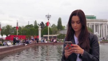 Parkta çekici, gülümseyen kız, fıskiyenin yanında oturuyor, akıllı telefon kullanıyor
