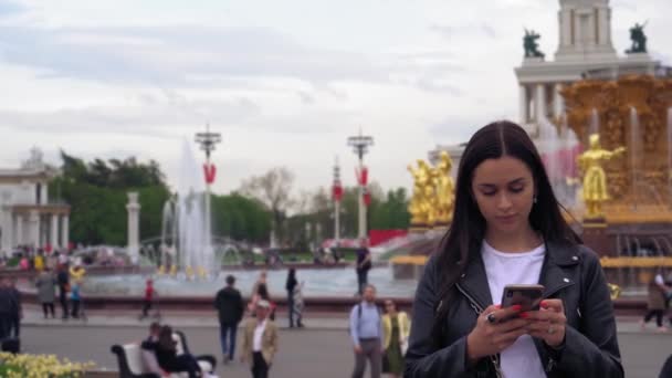 Vonzó fiatal nő egy parkban, gépelés egy mobil telefon, mosolyogva. Lassított mozgás — Stock videók