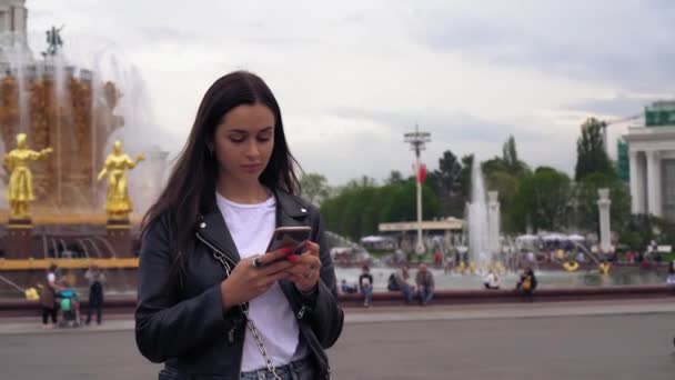 Aantrekkelijke jonge vrouw typen op een mobiele telefoon, kijkt rond, glimlachend. — Stockvideo