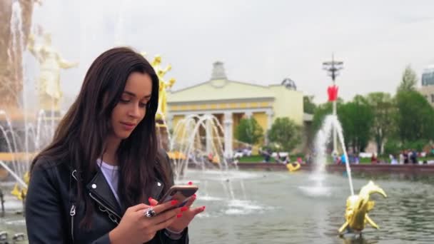 Vonzó fiatal nő sétál a parkban. Használ a mobiltelefonján. Stílusos viselet. — Stock videók
