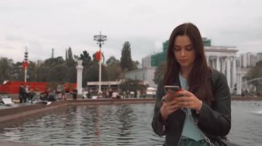 Bir ceket ve kot pantolon çekici kız, akıllı telefon ile çeşme yakınında oturan