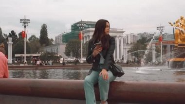 Bir ceket ve kot pantolon çekici kız, akıllı telefon ile çeşme yakınında oturan
