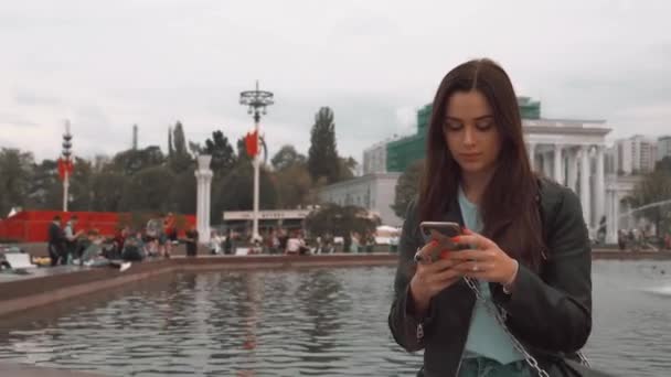 Bir ceket ve kot pantolon çekici kız, akıllı telefon ile çeşme yakınında oturan — Stok video