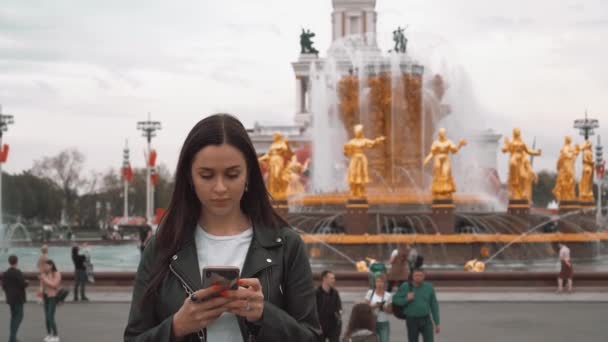 Menina agradável em jaqueta preta e t-shirt branca, digitando SMS no smartphone. Fechar — Vídeo de Stock