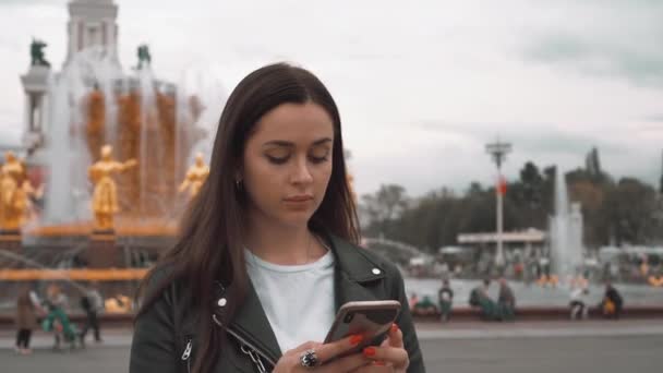 Menina agradável em jaqueta preta e t-shirt branca, digitando SMS no smartphone. Fechar — Vídeo de Stock