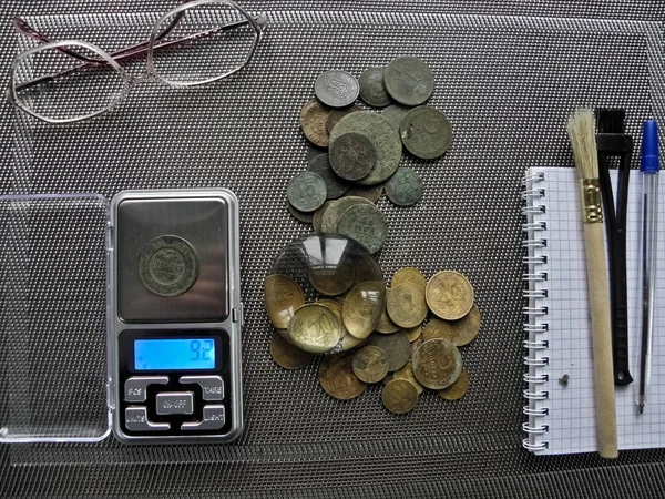 A lot of old copper coins for restoration, are lying on the table, on a light background, near the means for cleaning, scales, a candle, glasses, a notebook, a fountain pen, brushes, a magnifier.