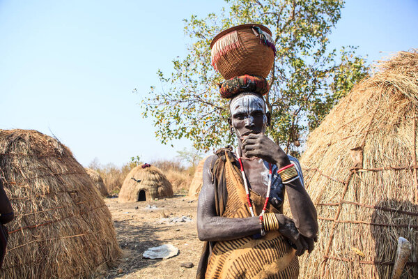Jinka, Omo River Valley, Ethiopia - January, 2018. Mursi Tribe man carrying a jar on his head, standing near hut, posing to tourists and covering his mouth with a hand