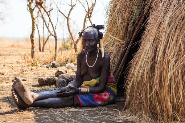 Jinka, Omo River Valley, Ethiopia - January, 2018. Mursi Tribe old woman sitting on ground at the entrance to her hut.