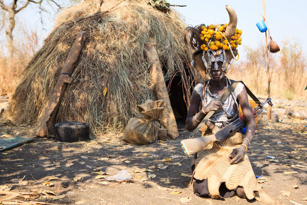 Jinka, Omo River Valley, Ethiopia - January, 2018. Mursi Tribe woman holding Kalashnikov gun, sitting in front of her hut, carrying lots of head decorations