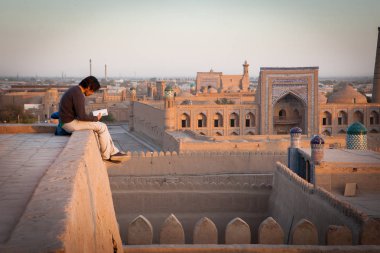 Khiva, Uzbekistan - October, 2016. A lonely tourist sitting on the roof of one of houses in Old City of Khiva, reading a book and enjoying romantic and picturesque sunset. clipart