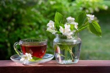 Bir fincan çay ve yasemin dallarıyla dolu bir vazo. Bokeh ile yeşil bulanık arka plan. Yaz havası. Parlak yaz güneşli gün. 