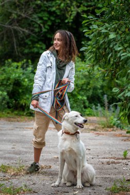 Dreadlocks ile Latino kız bir tasma bir köpek tutar. Yaz. Gree bulanık arka plan. Hayvanla iletişim. Yaz.