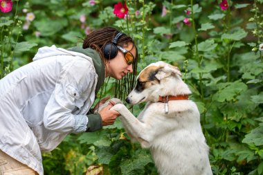 Çiçeklerin arka planında köpek köpeğinin yanındaki kulaklık ve gözlüklü kız. Beyaz bir ceket giyen dreadlocks ile görünüm Latino kız. Yaz günü. Hayvanla iletişim. 