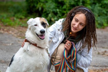 Bulanık yeşil arka planda köpek pooch yanındaki kulaklık kız. Beyaz bir ceket giyen dreadlocks ile görünüm Latino kız. Yaz günü. 