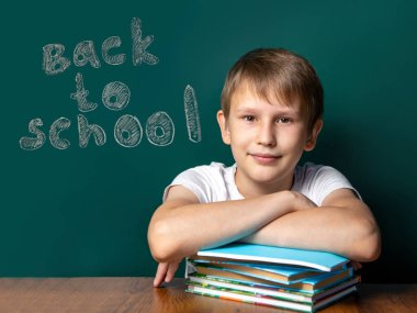 Okul kurulu arka planda Kafkas görünümü bir çocuk. Onun yanında defter ve ders kitapları yığını vardır. Kameraya bakıyorum. Yeşil arka plan. Okul yılı başlangıcı