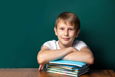 Okul kurulu arka planda Kafkas görünümü bir çocuk. Onun yanında defter ve ders kitapları yığını vardır. Kameraya bakıyorum. Yeşil arka plan. Okul yılı başlangıcı