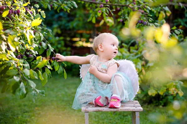 El bebé está sentado en el jardín. Chica ángel. Día soleado de verano. Árboles en el jardín. Fondo borroso verde en el fondo — Foto de Stock