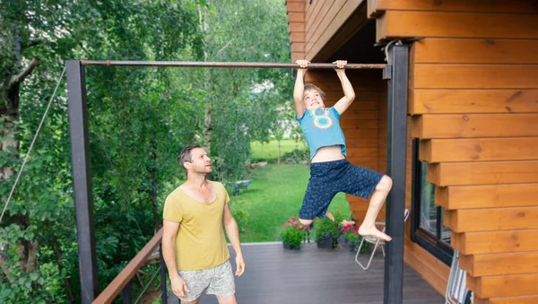 Achieving the goal through effort. The boy pulls himself up on the gymnastic bar. Sportive family doing exercises outdoors. The father teaches his son to pull himself up on the bar.