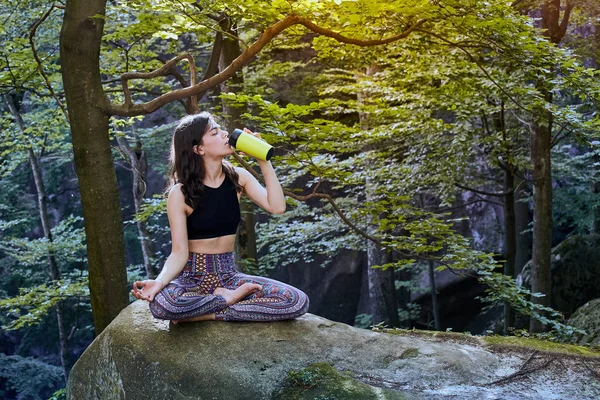 Sano fitness ragazza seduta in posa di loto e acqua potabile da bottiglia verde nella foresta — Foto Stock