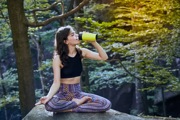 Здорова фітнес-дівчина сидить у позі лотоса і п'є воду з зеленої пляшки в лісі — стокове фото