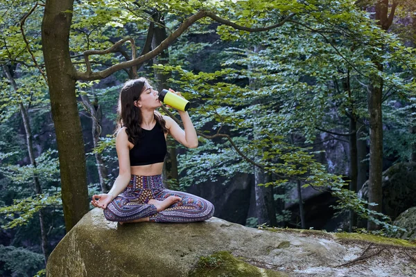 Здорова фітнес-дівчина сидить у позі лотоса і п'є воду з зеленої пляшки в лісі — стокове фото
