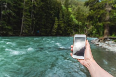 Yazın dağlarda güzel bir nehir ve ormanın arka planında telefonu olan bir kadının birinci şahıs görüntüsü. Tatil ve turizm konsepti