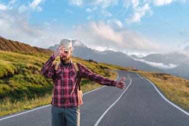 Hasır şapkalı ve ekose gömlekli yakışıklı genç adam, gün batımında güzel dağların zeminine karşı yolda duruyor. Özgürlük ve otostop konsepti