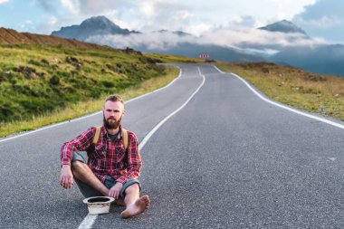 Ekose gömlekli ve şortlu yakışıklı genç adam gün batımında dağlarda bir patikada oturur. Özgürlük ve seyahat konsepti
