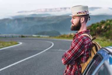 Hasır şapkalı ve ekose gömlekli yakışıklı genç adam, gün batımında güzel dağların zeminine karşı yolda duruyor. Özgürlük ve otostop konsepti