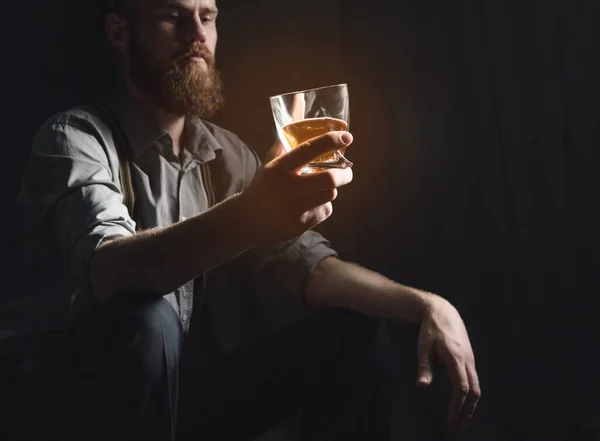 Um jovem bonito com barba está sentado em um quarto escuro com um copo de álcool em suas mãos. O conceito de descanso e relaxamento, bebida e solidão — Fotografia de Stock