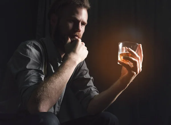 Красивый молодой человек с бородой сидит в темной комнате со стаканом алкоголя в руках. Концепция отдыха и релаксации, выпивки и одиночества — стоковое фото
