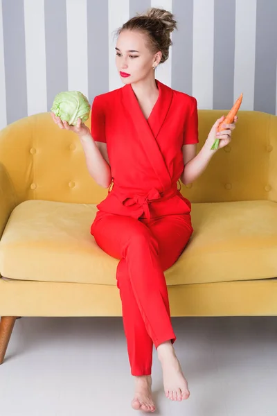 Uma linda garota em um macacão vermelho se senta em um sofá amarelo e segura vegetais em suas mãos. O conceito de um estilo de vida saudável e juventude — Fotografia de Stock