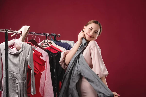 Menina bonita escolhe e experimenta roupas em um fundo vermelho. O conceito de escolher e comprar roupas, shopaholism e consumismo — Fotografia de Stock