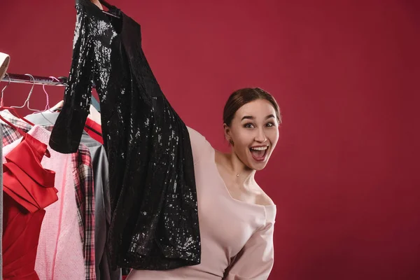 Menina bonita escolhe e experimenta roupas em um fundo vermelho. O conceito de escolher e comprar roupas, shopaholism e consumismo — Fotografia de Stock