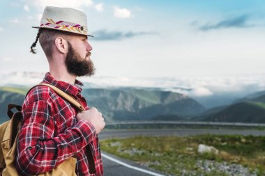 Hasır şapkalı ve ekose gömlekli yakışıklı genç adam, yolda duruyor, güzel dağların arka planında. Özgürlük ve otostop konsepti