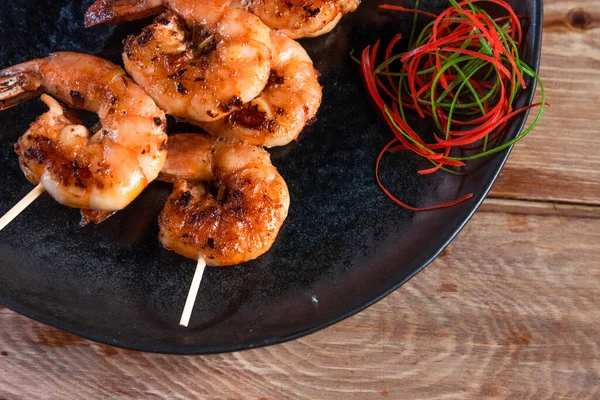 Apetitivo camarón frito en un pincho en un plato negro sobre una superficie de madera entre limón y condimentos. El concepto de comida rápida y saludable — Foto de Stock