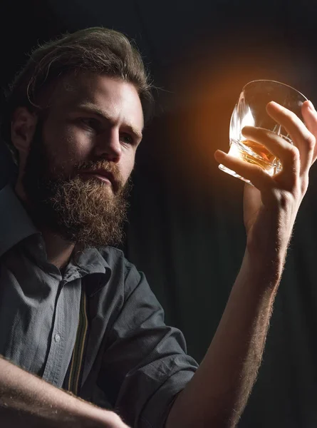 Um jovem bonito com barba está sentado em um quarto escuro com um copo de álcool em suas mãos. O conceito de descanso e relaxamento, bebida e solidão — Fotografia de Stock