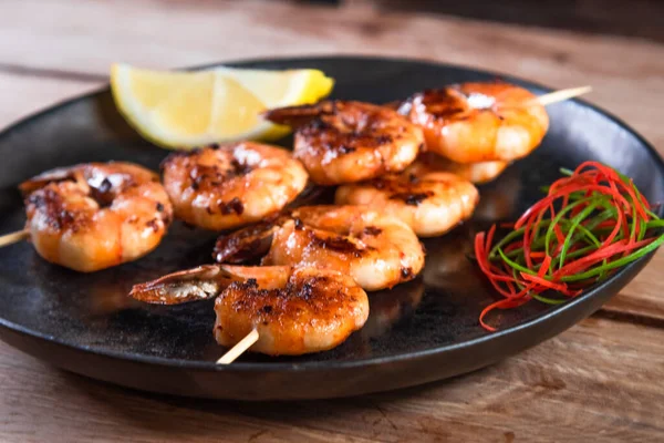 Apetitivo camarón frito en un pincho en un plato negro sobre una superficie de madera entre limón y condimentos. El concepto de comida rápida y saludable — Foto de Stock