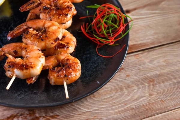 Apetitivo camarón frito en un pincho en un plato negro sobre una superficie de madera entre limón y condimentos. El concepto de comida rápida y saludable — Foto de Stock