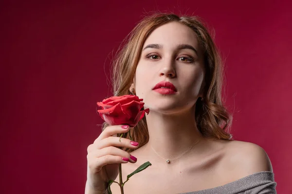 Menina bonita com maquiagem brilhante, segurando uma rosa em suas mãos em um fundo vermelho no estúdio. O conceito de juventude, beleza e um corpo saudável — Fotografia de Stock