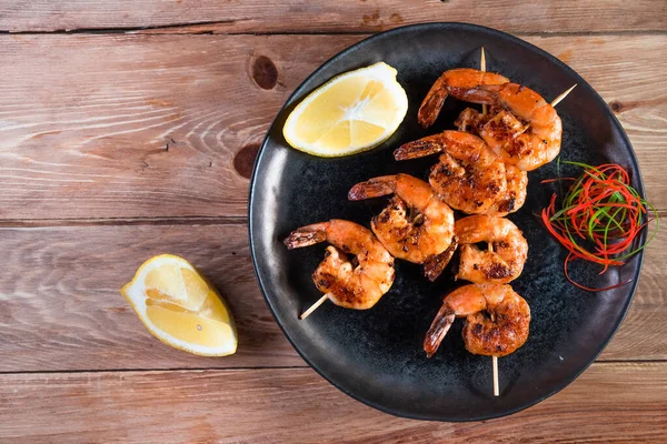 Apetitivo camarón frito en un pincho en un plato negro sobre una superficie de madera entre limón y condimentos. El concepto de comida rápida y saludable — Foto de Stock