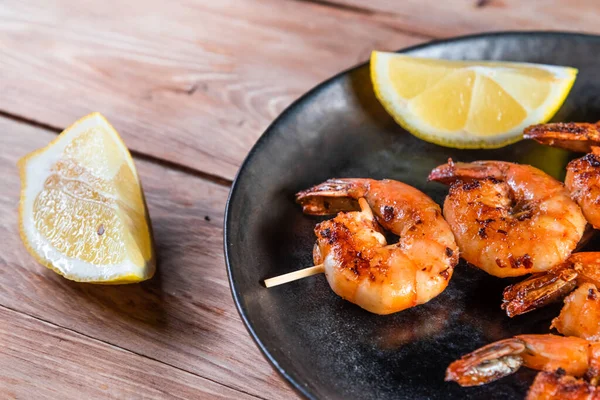 Apetitivo camarón frito en un pincho en un plato negro sobre una superficie de madera entre limón y condimentos. El concepto de comida rápida y saludable — Foto de Stock