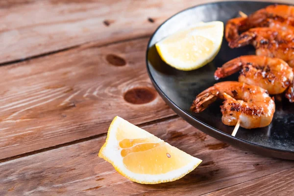 Apetitivo camarón frito en un pincho en un plato negro sobre una superficie de madera entre limón y condimentos. El concepto de comida rápida y saludable — Foto de Stock