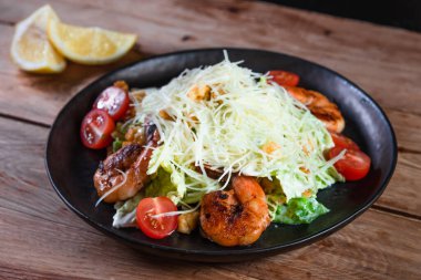 İştah açıcı Sezar salatası, siyah bir tabakta karides, ahşap bir masada, limonun yanında. Sağlıklı yemek ve hizmet kavramı