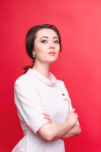 Trabalhadora atraente do campo médico em uniforme fica no estúdio contra o pano de fundo de uma parede vermelha. O conceito de medicina, cosmetologia e massagem — Fotografia de Stock