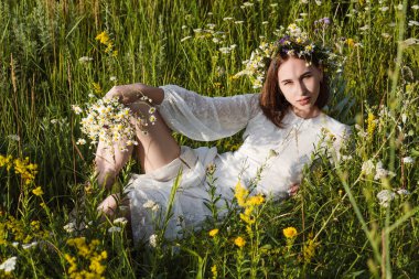 Beyaz elbiseli, başında çelenk olan güzel bir kadın çiçeklerle bir tarlada yatıyor. Güzellik, özgür yaşam ve doğallık kavramı.