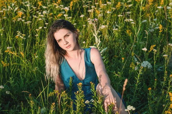 Uma mulher loira bonita em um vestido longo azul que se senta em um campo em flores. O conceito de beleza, vida livre e naturalidade — Fotografia de Stock